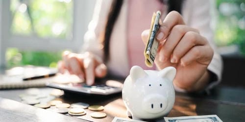 A person inserting cash into a piggy bank while using a calculator, with coins and bills spread on the table.