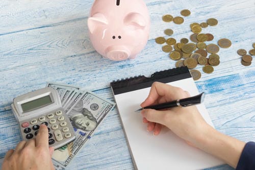 Hands budgeting with a calculator, notepad, cash, coins, and a pink piggy bank on a light blue wooden table.