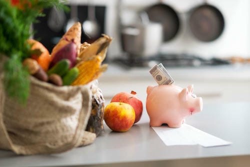 A kitchen counter scene with a reusable grocery bag of produce, apples, and a piggy bank with cash and a receipt.