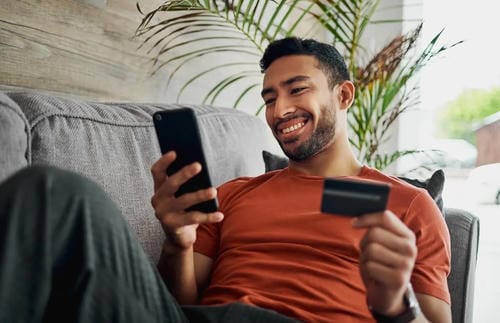A smiling man on a couch using a smartphone while holding a credit card for online payments