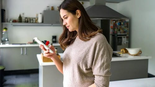 A woman using her smartphone in a modern kitchen, likely managing her finances or banking.