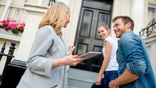 A real estate agent using a tablet to show a property to a young couple outside a house.