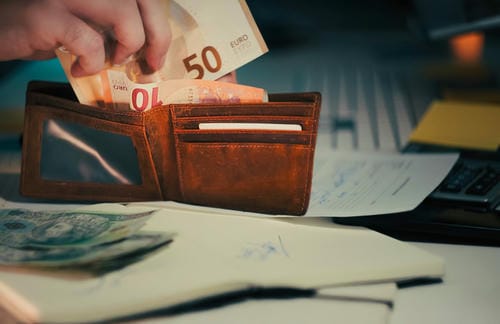A hand placing 10 and 50 Euro banknotes into a brown leather wallet, with financial documents nearby.