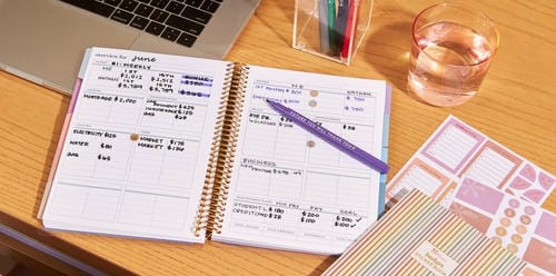 An open budget planner with handwritten income and expense entries, alongside a laptop and stickers.
