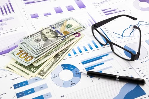 US dollar bills resting on financial documents with charts, a pen, and glasses, illustrating financial analysis.