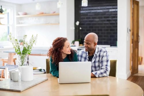 A couple smiling and looking at each other while using a laptop at home, likely discussing finances or plans.