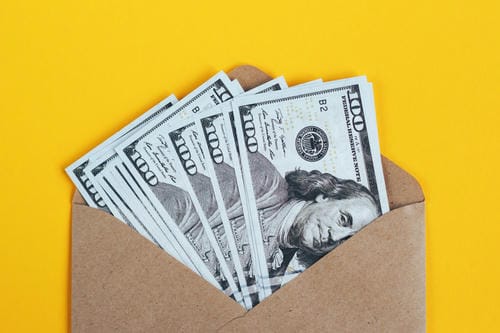US $100 bills fanned out inside a brown envelope against a bright yellow background.