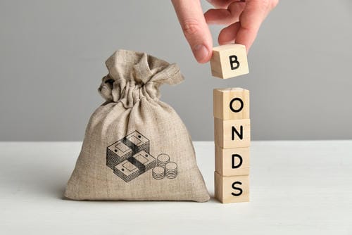 A hand placing a wooden block letter "B" to spell "BONDS" next to a money bag, symbolizing bond investments.