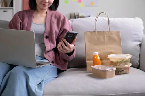 A woman uses her phone and laptop while ordering takeout food, a common example of everyday spending.