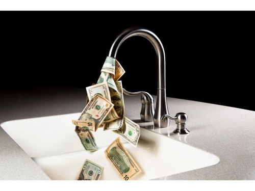 US dollar bills flow from a kitchen faucet into the sink, visually representing the idiom “money down the drain” or wasting money.