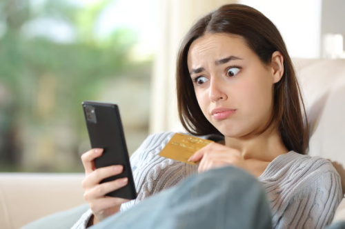 A woman looks concerned while holding a credit card and checking her smartphone, likely reacting to an unexpected expense or bill.
