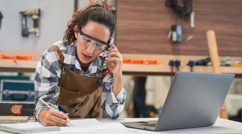 A woman carpenter in safety glasses, apron, and plaid shirt talks on the phone while writing notes and using a laptop in her workshop.