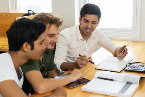 Three diverse students studying together at a wooden table with notebooks, a calculator, and a tablet.